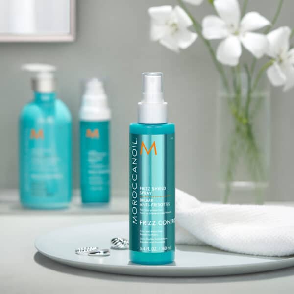 Frizz Shield Spray de Moroccanoil en baño sobre plato blanco con fondo de flores.