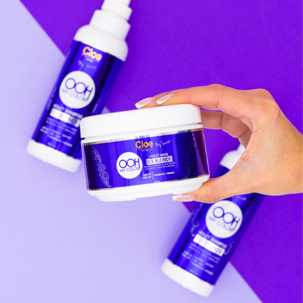 Mano sosteniendo envase de mascarilla Cloe OOH My Color Violet Mask Ice Blonde, con botellas de shampoo desenfocadas al fondo sobre superficie morada.
