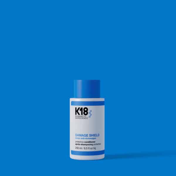 El K18 Damage Shield Acondicionador de 250 ml está de pie sobre un fondo azul sólido. Su envase es blanco con una franja azul en la parte superior y un tapón azul brillante. La etiqueta muestra el logo K18 Biomimetic Hairscience en negro, con detalles del producto en azul y negro. Este acondicionador profesional protege el cabello de agresores externos y ayuda a mantener su hidratación.