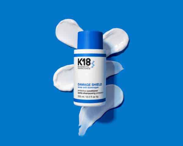 El K18 Damage Shield Acondicionador de 250 ml está colocado sobre un fondo azul con varias muestras de su textura cremosa de color blanco esparcidas a su alrededor. Su envase es blanco con una franja azul y un tapón azul brillante. La etiqueta resalta el logo K18 Biomimetic Hairscience, y el acondicionador está formulado para suavizar, proteger y fortalecer la fibra capilar.