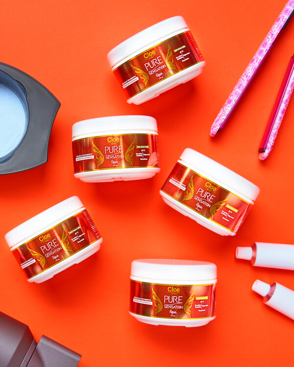 Cinco envases de mascarilla capilar Cloe Pure Sensation Reparación dispuestos sobre fondo rojo. Alrededor se observan utensilios de peluquería como plancha y bowl.