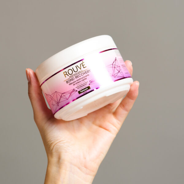 Mano sosteniendo envase de mascarilla Rouvé Bond Recovery de 270 gramos, con fondo gris claro.