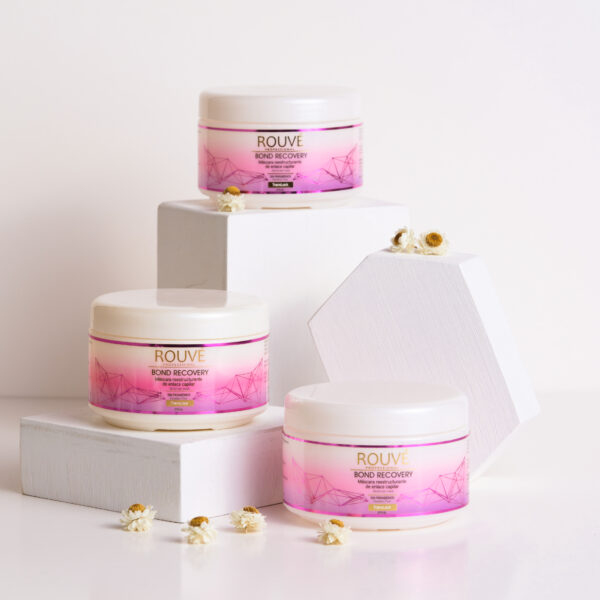 Tres envases de mascarilla Rouvé Bond Recovery dispuestos sobre bases blancas de diferentes alturas, decorados con pequeñas flores blancas y fondo blanco.