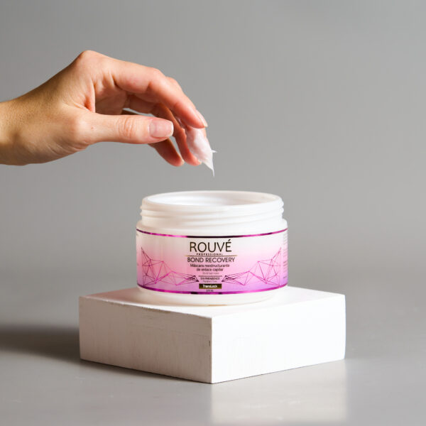 nvase abierto de mascarilla Rouvé Bond Recovery sobre base blanca, con una mano tomando el producto cremoso y fondo gris claro.