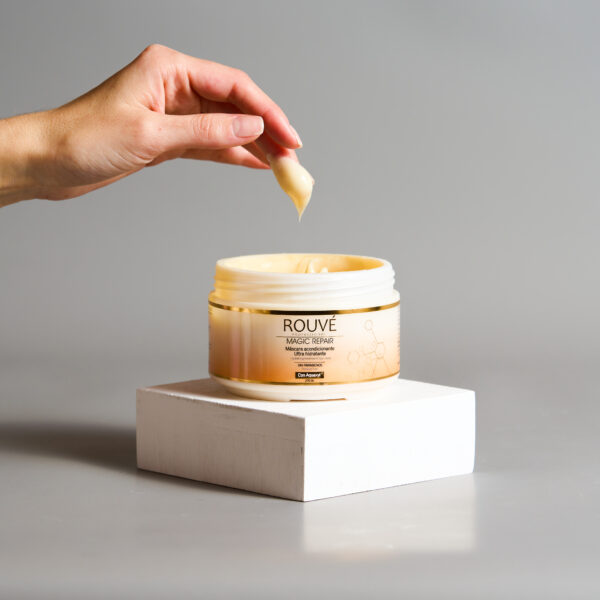 Envase abierto de mascarilla Rouvé Magic Repair sobre base blanca, con una mano tomando producto y fondo gris claro.